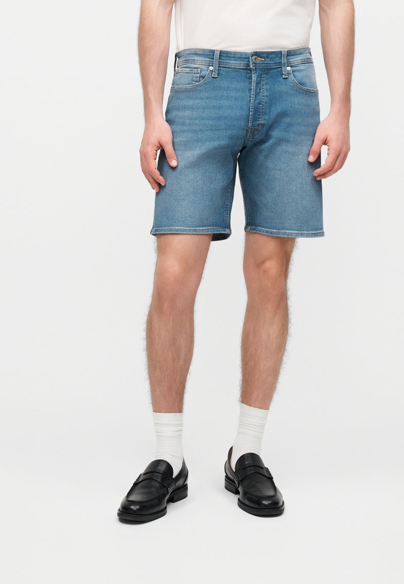 Άνδρας που φοράει μεσαίου μπλε τζιν σορτς, λευκές αθλητικές κάλτσες, μαύρα μοκασίνια και λευκό μπλουζάκι, στέκεται μπροστά σε απλό φόντο.