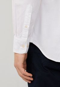 Camicia bianca con colletto a punta, caratterizzata da due bottoni in perla. Il tessuto sembra liscio e leggermente testurizzato, infilato in pantaloni scuri.