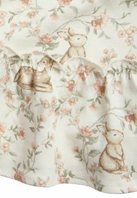 Blommönstrad tyg med beige kaniner och rosa blommor på en off-white bakgrund. Inkluderar volangdetaljer längs kanten.