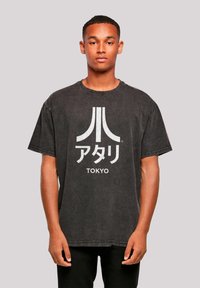 Schwarzes Oversized-T-Shirt aus Baumwolle, mit einem auffälligen weißen Atari-Logo und dem japanischen Text "TOKYO", der auf der Vorderseite aufgedruckt ist.