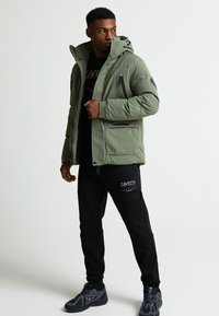 Olīvu zaļa pufīgā jaka ar kapuci, ar rāvējslēdzēja kabatām un kontrastējošām melnām akcentiem; apvienota ar melnām sporta biksēm ar logotipu.