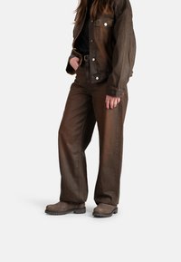 Chaqueta de mezclilla marrón y pantalones de pierna ancha a juego, ambos con un acabado texturizado. La modelo lleva botas gris de slip-on con una suela resistente.