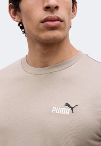 Νέος άνδρας που φοράει μπεζ μπλουζάκι Puma με στρογγυλή λαιμόκοψη και μικρό λογότυπο Puma στο στήθος.