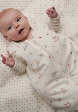 Baby med vidåpne øyne liggende på et blekt pledd med polkaprikkmønster, iført en hvit sovepose dekorert med små leker og klossemønster.