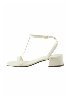 Vit vegansk läder sandal med T-rem design, fyrkantig tå och en texturerad blockklack med ett subtilt mönster. Guldspänne som accent.