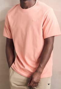 Camiseta de algodón rosa con mangas cortas y escote redondo. Presenta un pequeño logo negro de Nike en el pecho. Llevada con pantalones beige.