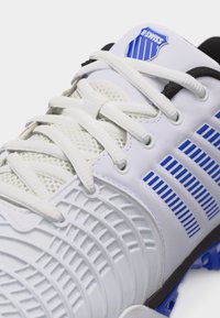 Άσπρο αθλητικό παπούτσι K-Swiss με μπλε ρίγες και λογότυπο, λευκά κορδόνια και υφή στην σόλα, εμφανίζεται σε κοντινή λεπτομέρεια.