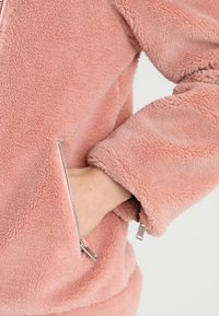 Różowa polarowa kurtka o miękkiej, teksturowanej powierzchni. Posiada zamykaną na zamek kieszeń oraz luźny krój z szerokimi rękawami.