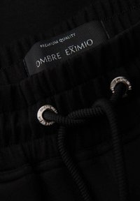 Czarne dresy z sznurkiem ze srebrnymi elementami, pasem z materiału prążkowanego oraz metką z napisem "OMBRE EXIMIO."