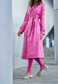 Trench coat lungo di un vivace rosa, con vita regolabile e bottoni decorativi, abbinato a leggings rosa e scarpe con zeppa in stampa leopardata.