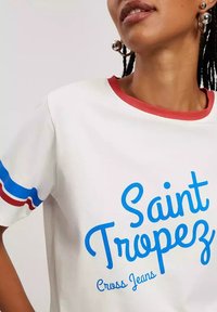 Biały t-shirt z krótkim rękawem z czerwonymi i niebieskimi paskami na rękawach, z napisem "Saint Tropez" w niebieskim kursywie.