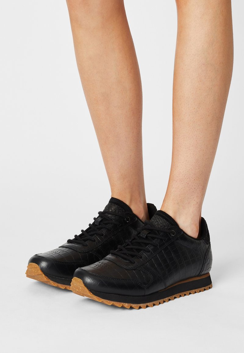 Personne portant des baskets basses noires en cuir texturé avec des semelles en caoutchouc gomme sur un fond blanc.