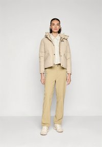 Veste matelassée beige avec capuche, dotée de boutons-pression et de poches latérales, portée sur une chemise claire et un pantalon beige. Le mannequin pose devant un fond neutre.