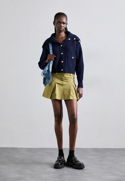 Navy wool blend jacket with large buttons and front pockets, paired with a pleated olive green skirt and chunky black loafers. Light blue crossbody bag.