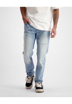 Personne portant un jean bleu clair déchiré, un t-shirt blanc et des baskets noires, marchant une main dans la poche sur un fond uni.