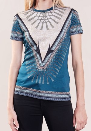 Femme portant une chemise ajustée à manches courtes bleue avec des motifs géométriques complexes et des motifs en forme de pompons, associée à un pantalon noir.