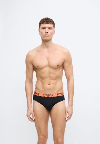 Άνδρας με κοντά σγουρά μαλλιά στέκεται προς τα εμπρός φορώντας μαύρα σλιπ με πορτοκαλί μέση που φέρει το λογότυπο EA σε απλό φωτεινό φόντο.