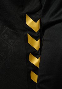 Schwarzer Stoff mit einem subtilen gemusterten Hintergrund und einem kräftigen gelben Zickzackdesign an der Seite. Weiche Textur, sportlicher Schnitt.