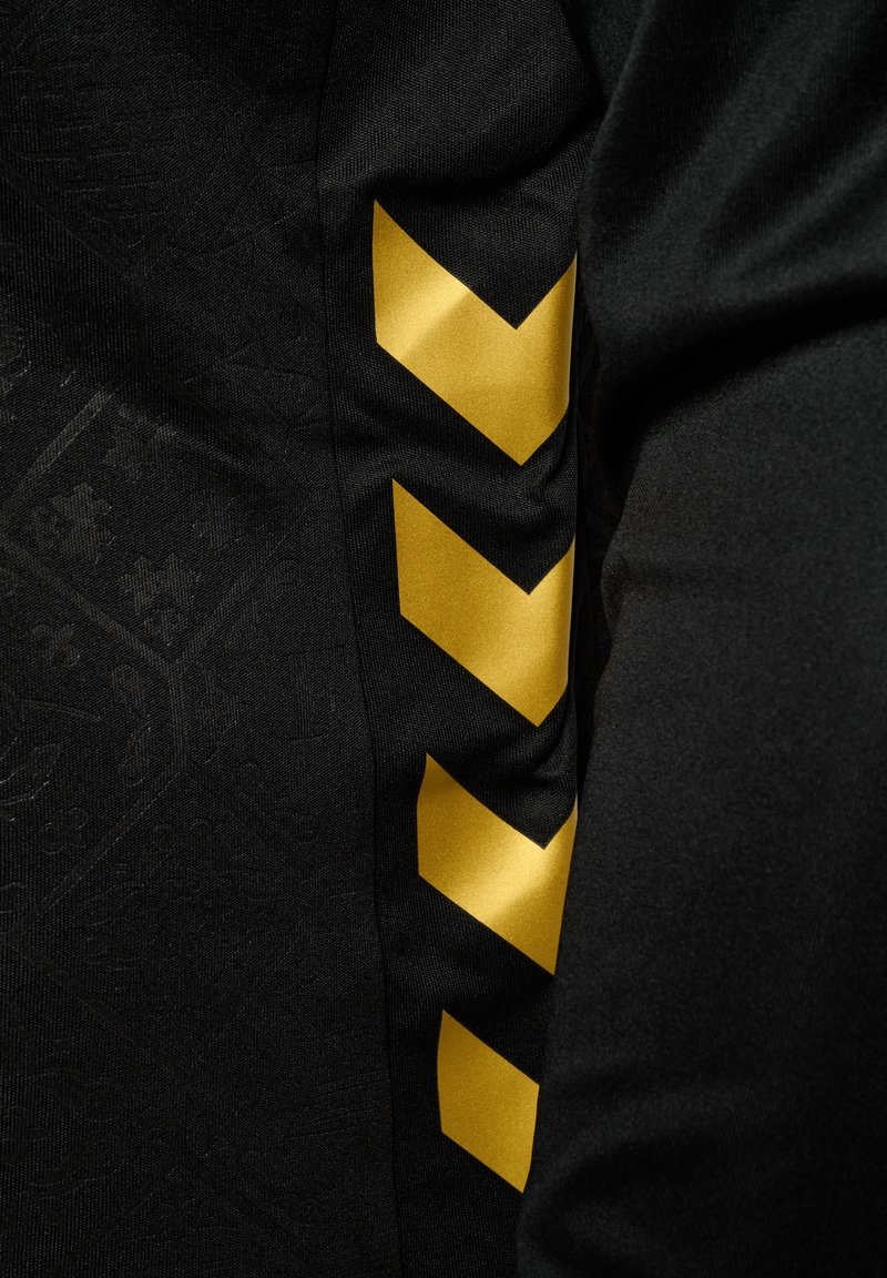 Schwarzer Stoff mit einem subtilen gemusterten Hintergrund und einem kräftigen gelben Zickzackdesign an der Seite. Weiche Textur, sportlicher Schnitt.