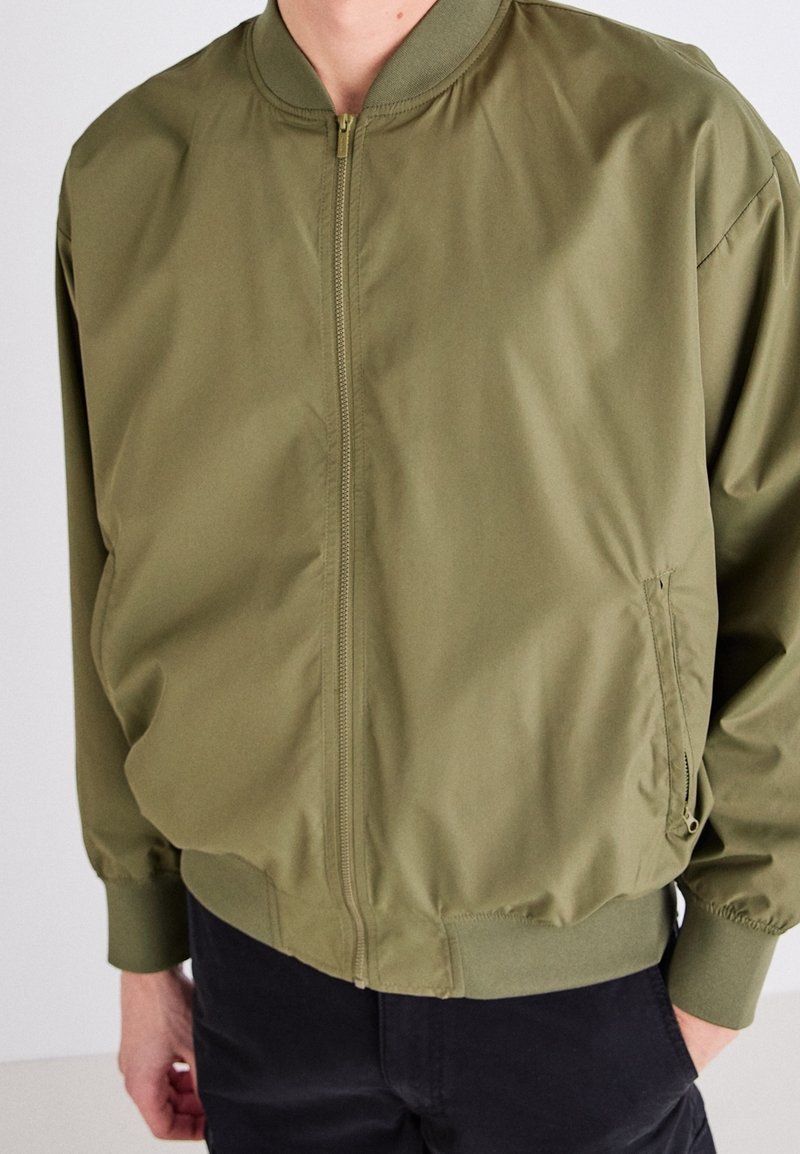 Giacca bomber verde oliva con zip frontale, colletto e polsini a coste. Realizzata in tessuto liscio e leggero con tasche laterali.