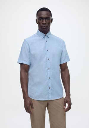 Man met een lichtblauw kortärmelig overhemd met knopen en beige broek, staand tegen een effen witte achtergrond.