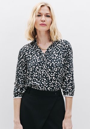 Blouse à pois noir et blanc avec des manches longues, col à boutons, plastron avant et une texture lisse, associée à une jupe noire.