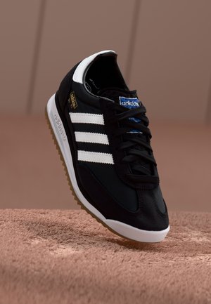 Baskets noires Adidas avec des bandes blanches et un détail au talon, logo bleu sur la languette, reposant sur une surface marron texturée.