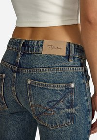 Világoskék farmernadrág magas derékkal, bézs bőrpöttyökkel és díszvarrással a hátsó zsebein.