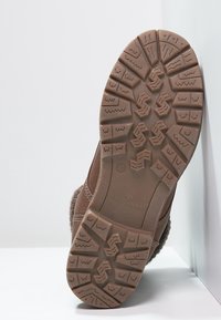 Hnědá obuv s texturovanou gumovou podrážkou s výrazným vzorem, šitými švy a měkkým, chlupatým vrchním materiálem.