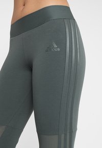 Leggings grigi alti in vita realizzati in materiale elastico, con tre strisce verticali e un logo in rilievo sulla coscia.