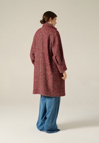 Cappotto in misto lana di color bordeaux con un motivo a spina di pesce testurizzato. Presenta un colletto alto e maniche ampie. Abbinato a jeans larghi blu chiaro.