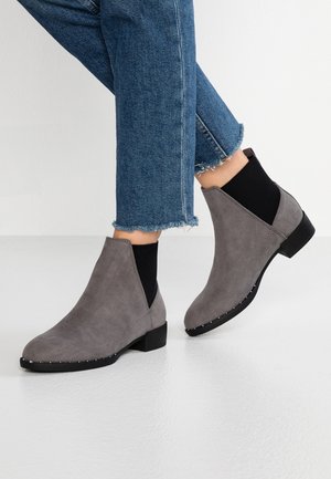 Persona che indossa jeans blu corti e stivaletti in camoscio grigio con pannelli elastici neri e tacchi bassi a blocchetto su un pavimento bianco.