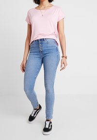 Camiseta de manga corta rosa claro combinada con jeans ajustados de cintura alta en azul con cierre de botones, y zapatillas negras con detalles en blanco.