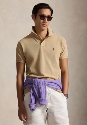 Muž nosiaci béžové polotričko, biele šortky, fialový sveter zviazaný okolo pásu, slnečné okuliare a náramkové hodinky, stojaci pred neutrálnym pozadím.