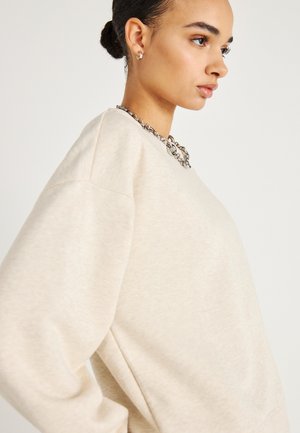 Felpa color beige chiaro con maniche lunghe e morbide e scollo arrotondato. Presenta una collana di catena d'argento come accessorio distintivo.