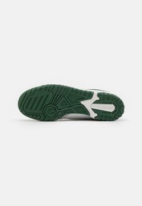 Suola di sneaker bianca con battistrada in gomma verde, caratterizzata da scanalature orizzontali e verticali e il nome del marchio "new balance" impresso sul lato.