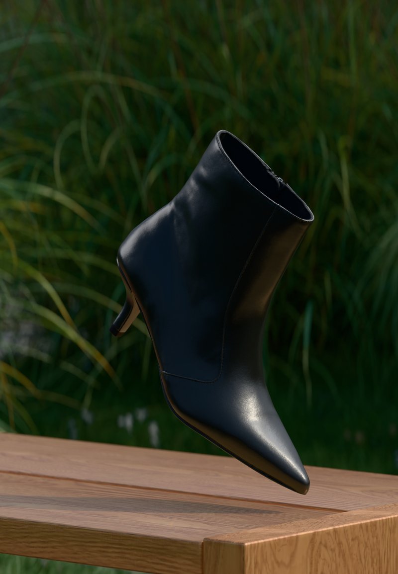 Čierne kožené členkové čižmy s ostrou špičkou a stredne vysokým kitten podpätkom levitujúce nad dreveným povrchom na pozadí zelenej listnatiny.