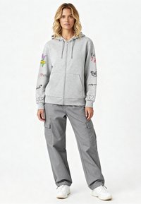 Szürke cipzáras kapucnis pulóver színes hímzett mintákkal az ujjain, laza szürke cargo nadrággal és fehér tornacipővel párosítva.