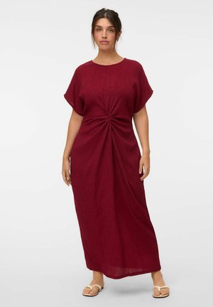 Mujer que lleva un vestido largo de manga corta y color rojo oscuro con un detalle de nudo recogido en la cintura, de pie con expresión neutral y sandalias.