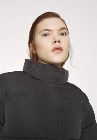 Μαύρο μπουφάν πούφ με ψηλό γιακά, που διαθέτει κουμπιά κλεισίματος και ορατές ραφές, φτιαγμένο από ομαλό, αδιάβροχο υλικό.