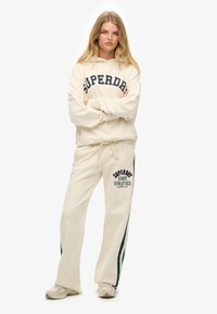 Beżowa bluza z kapturem z logo "SUPERDRY" oraz pasujące spodnie dresowe z napisem "SUPERDRY STATE ATHLETICS", obie z miękkiej, teksturowanej tkaniny.