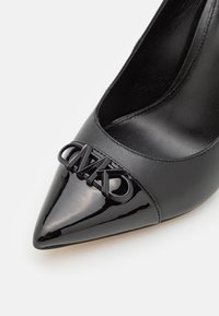 Gros plan d’un talon aiguilles noir à bout pointu présentant une coque brillante et un logo en relief à l’avant.