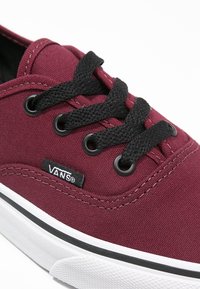 Burgunderfarbene Leinenschuhe mit schwarzen Schnürsenkeln, runden Ösen und weißer Gummisohle. Auf der Seite befindet sich ein kleines schwarzes Vans-Logo-Tag.