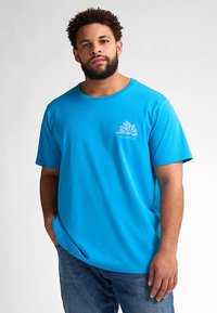 Modré bavlněné tričko s krátkými rukávy, kulatým výstřihem a malým potištěným logem na hrudi. Nošeno s modrými džínami.