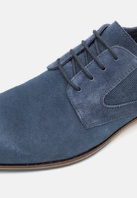 Zapato de vestir azul de ante con seis ojetes, panel lateral texturizado y acabado suave. Presenta una suela marrón clara y cordones grises.