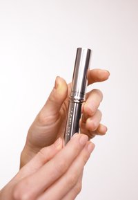 En metallisk sølvfarget maskara-tube holdt mellom fingrene, med en glatt, blank overflate og en avsmalnende design, som har subtilt gravert merkevare.