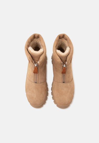 Paire de bottines en suède beige avec fermetures éclair à l’avant et doublure en fausse fourrure blanche moelleuse, vue de dessus sur fond blanc.