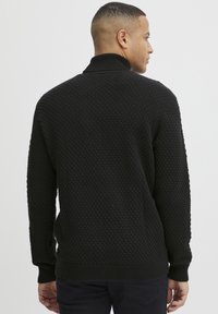 Maglione nero a maglia con superficie testurizzata, dotato di un colletto alto a coste e polsini a coste, mostrato da dietro, con una forma aderente.