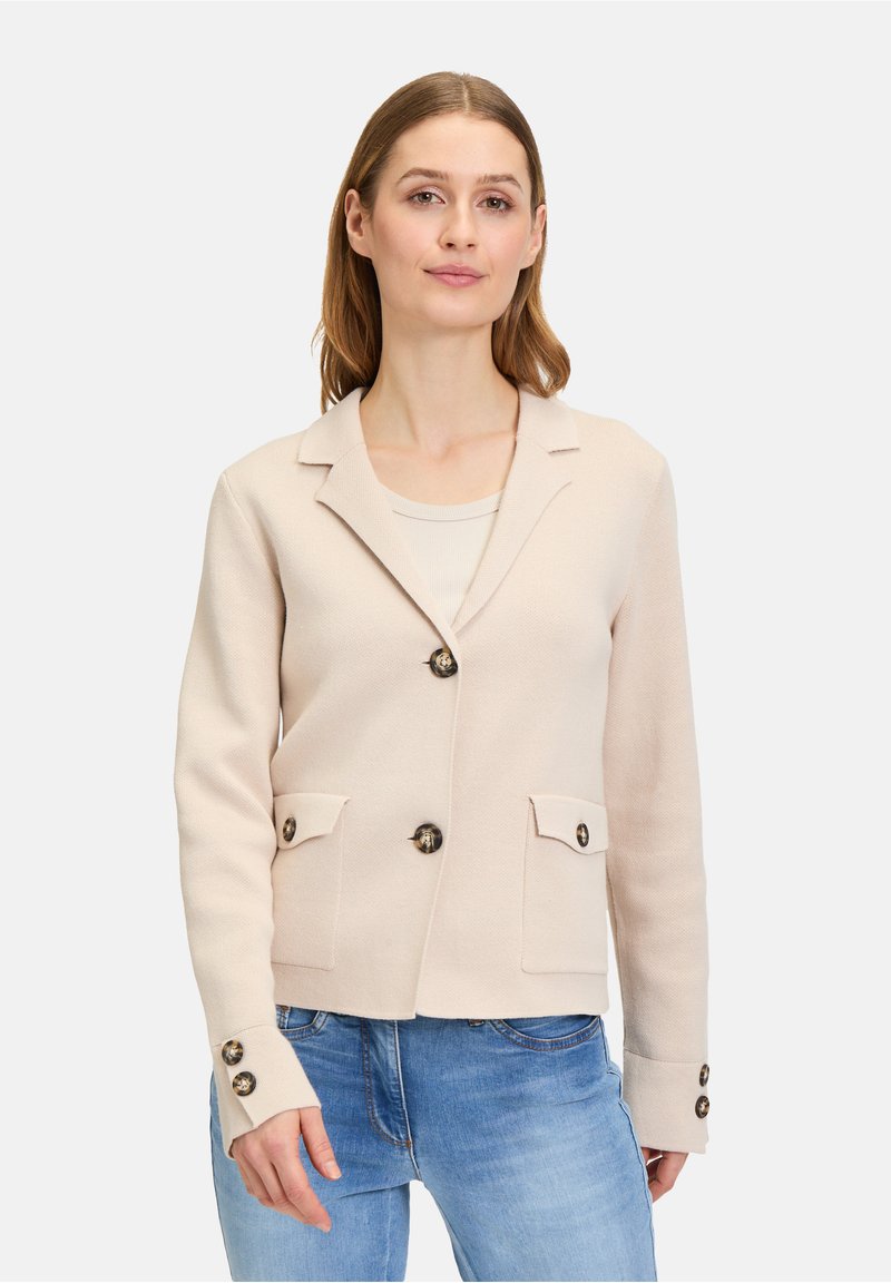 Blazer beige clair au design ajusté, avec deux poches avant et des boutons noirs, porté par-dessus un haut simple.