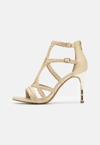 Beige korkokenkä korkokengät, joissa on punotut hihnat, avoin varvas ja takasuljenta. Metallinen yksityiskohta kannassa, sileä keinonahkamateriaali.